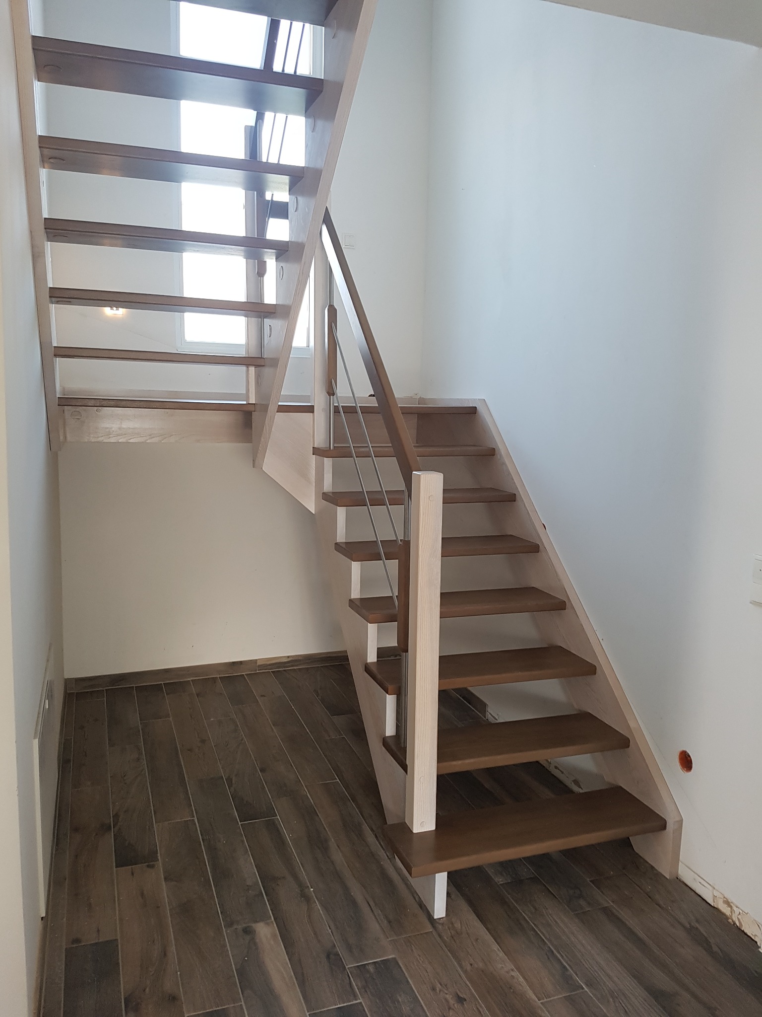 Drewniane schody z metalową balustradą w nowoczesnym wnętrzu, widoczne stopnie w kolorze ciemnego brązu i jasne drewniane elementy konstrukcyjne, na podłodze płytki imitujące drewno