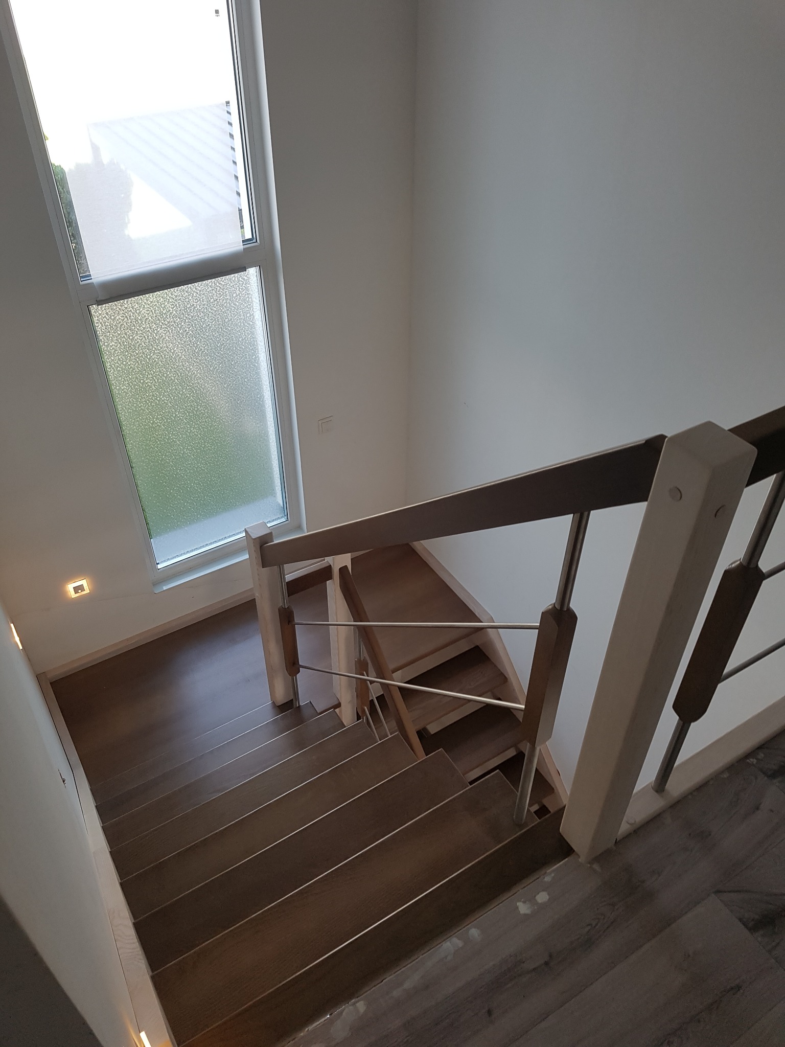 Widok z góry na nowoczesne schody z drewnianymi stopniami i balustradą z elementami drewna i stali nierdzewnej, oświetlone światłem z okna dachowego z matową szybą.