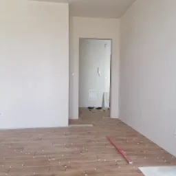 Układanie brązowych płytek imitujących drewno w jasnym pomieszczeniu, poziomica na podłodze, widoczne krzyżyki dystansowe.