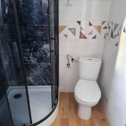 Nowoczesna łazienka z kabiną prysznicową z czarnymi profilami i marmurowym wykończeniem, białą toaletą oraz geometrycznym wzorem płytek na ścianie.