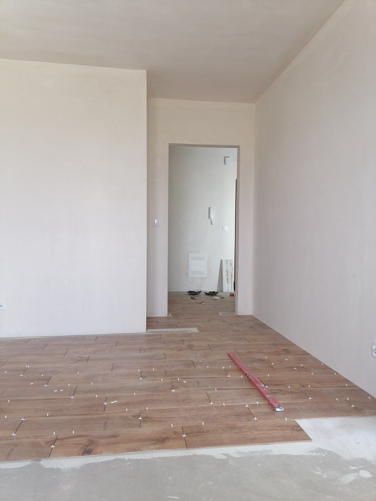 Układanie brązowych płytek imitujących drewno w jasnym pomieszczeniu, poziomica na podłodze, widoczne krzyżyki dystansowe.