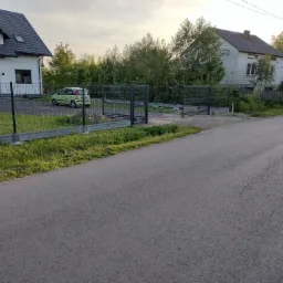 Nowoczesne, grafitowe ogrodzenie panelowe z podmurówką betonową i otwartą bramą wjazdową, widoczne z ulicy, z zielonym samochodem w tle.