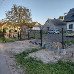 Z.K.weld - Montaż czarnej siatki ogrodzeniowej na betonowej podmurówce w wiejskiej scenerii, widoczne dwa samochody i budynki mieszkalne w tle.