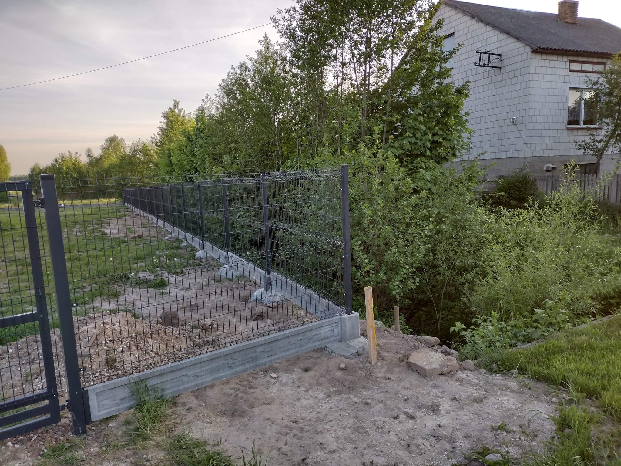 Fragment nowo postawionego, czarnego ogrodzenia z siatki na betonowej podmurówce, z widoczną bramką wejściową, w tle dom i zieleń.