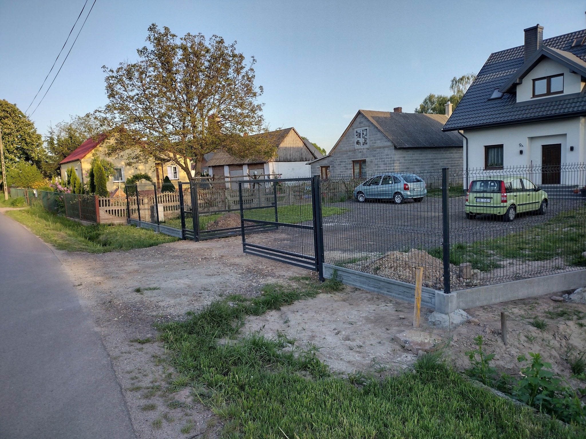 Montaż czarnej siatki ogrodzeniowej na betonowej podmurówce w wiejskiej scenerii, widoczne dwa samochody i budynki mieszkalne w tle.