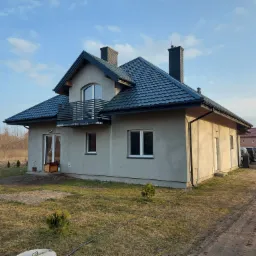 Nowoczesny dom jednorodzinny z ciemnoszarą dachówką, widoczny balkon z metalową balustradą, elewacja w trakcie wykończenia, teren wokół domu częściowo zagospodarowany.