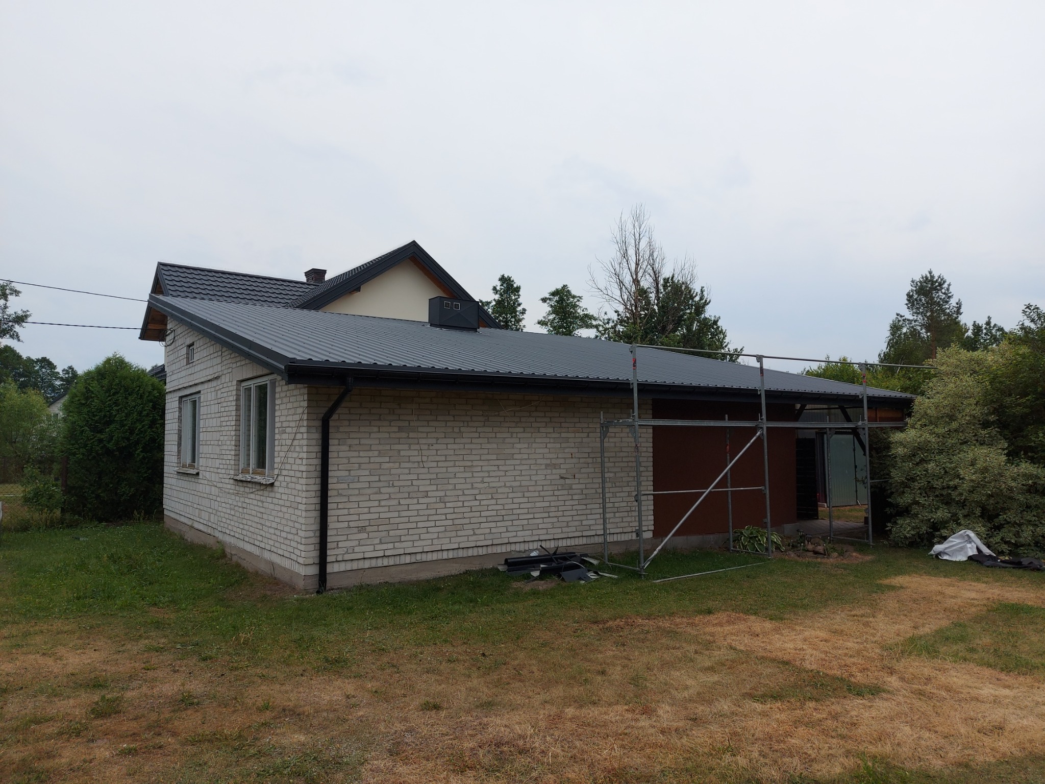 Dom z białej cegły z nowym, grafitowym dachem blaszanym, częściowo otoczony rusztowaniem, na trawniku widoczne elementy blacharskie.