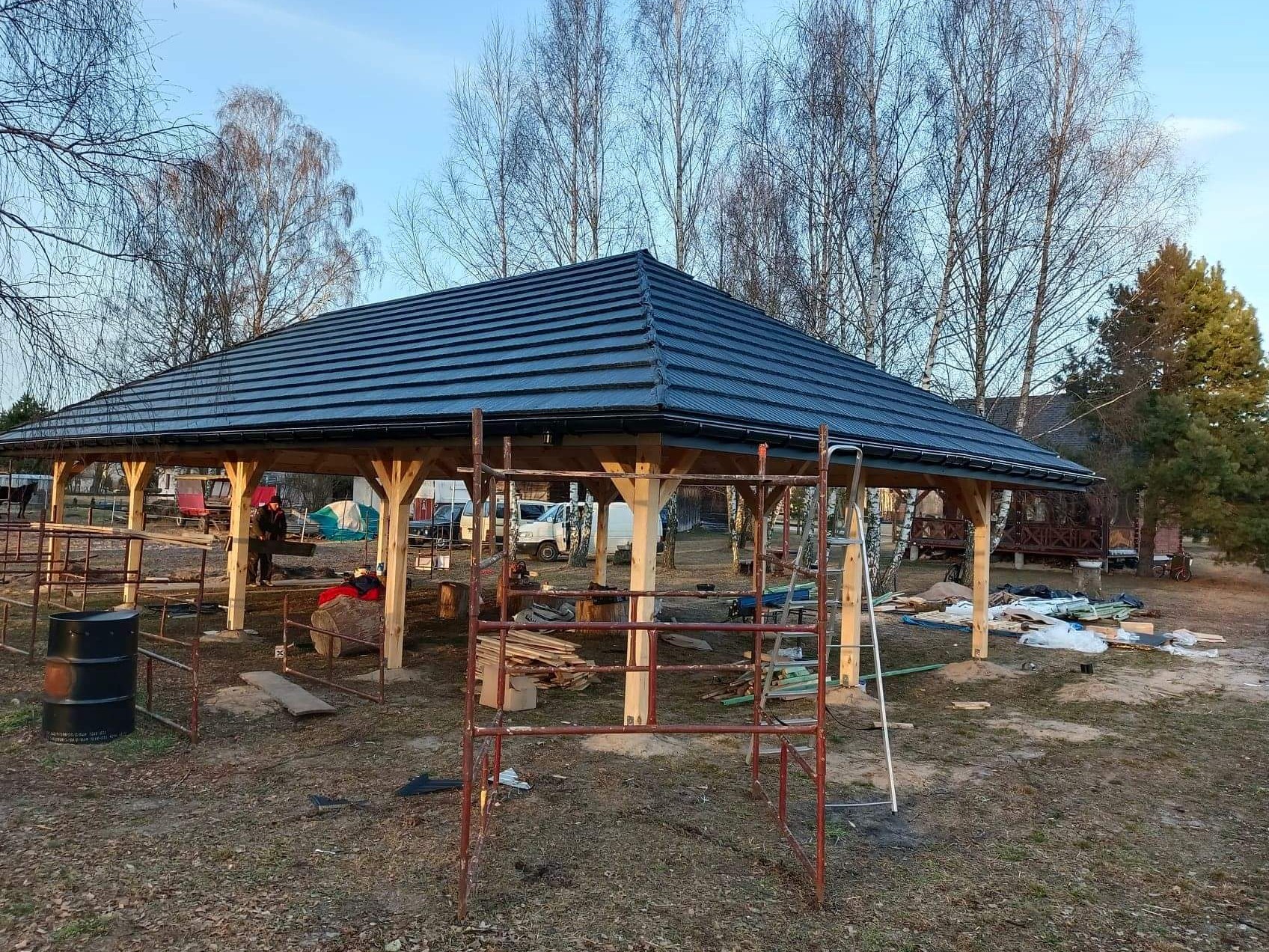 Montaż dachu z ciemnej blachodachówki na drewnianej konstrukcji altany ogrodowej, widoczne rusztowanie i drabina, w tle drzewa i zabudowania.