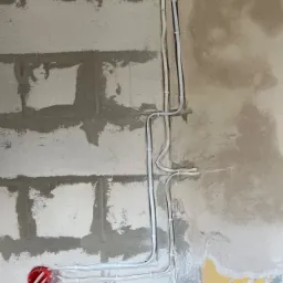 Surowa ściana z betonu komórkowego, częściowo pokryta resztkami starej farby i zaprawą, z widocznymi przewodami elektrycznymi w białych rurkach i czerwonym gniazdkiem w puszce.