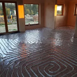 Rozłożone rury ogrzewania podłogowego na folii odblaskowej w surowym wnętrzu domu w budowie; w tle widoczne okna z widokiem na teren budowy i koparkę.