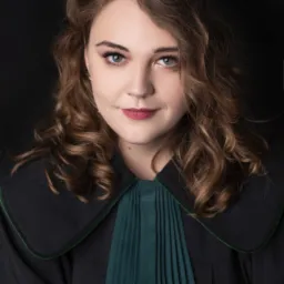 Portret kobiety-adwokata w todze na czarnym tle, z delikatnym uśmiechem i zielonymi oczami. Widoczne staranne upięcie włosów i subtelny makijaż.