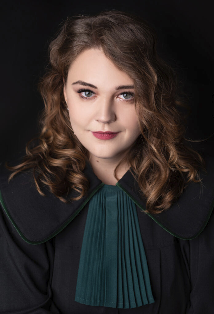 Portret kobiety-adwokata w todze na czarnym tle, z delikatnym uśmiechem i zielonymi oczami. Widoczne staranne upięcie włosów i subtelny makijaż.