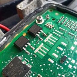 Zielona płytka drukowana z elementami elektronicznymi, widoczne układy scalone, tranzystory i ślady lutowania, prawdopodobnie element sterownika samochodowego.