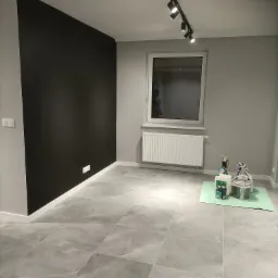 Pomalowane na czarno fragment ściany w pokoju z szarą podłogą z płytek. Widoczne puszki z farbą i akcesoria malarskie przygotowane na podłodze, grzejnik pod oknem i oświetlenie sufitowe.