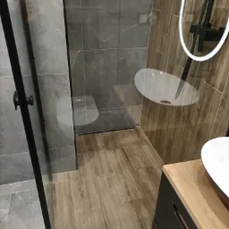 Nowoczesna łazienka po remoncie w Słupcy: kabina prysznicowa typu walk-in z czarnymi profilami, drewnopodobne płytki na podłodze, szare płytki na ścianach, umywalka nablatowa z szafką w kolorze...