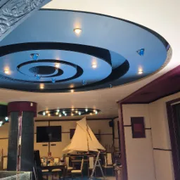 Malowanie sufitu w restauracji z ozdobnymi elementami i oświetleniem, widoczne zabezpieczenia podłogi folią malarską i drabina.