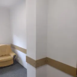 Fragment wnętrza biura lub mieszkania z białymi ścianami i szarym dywanem, z zamontowanymi drewnianymi listwami ochronnymi na ścianach, widoczny fragment żółtej kanapy