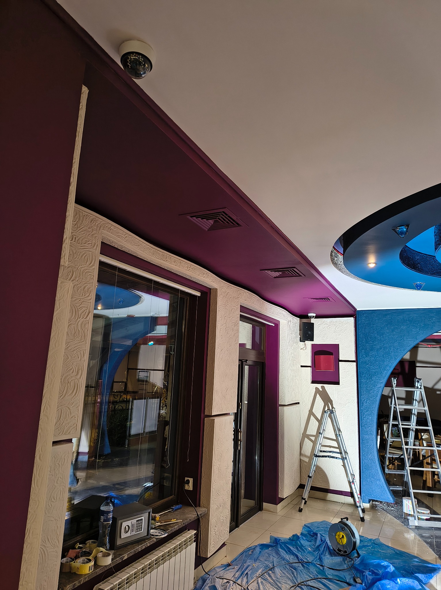 Wnętrze lokalu w trakcie malowania: fioletowe ściany i sufit, zabezpieczona folią podłoga, drabiny, widoczny sejf na parapecie.