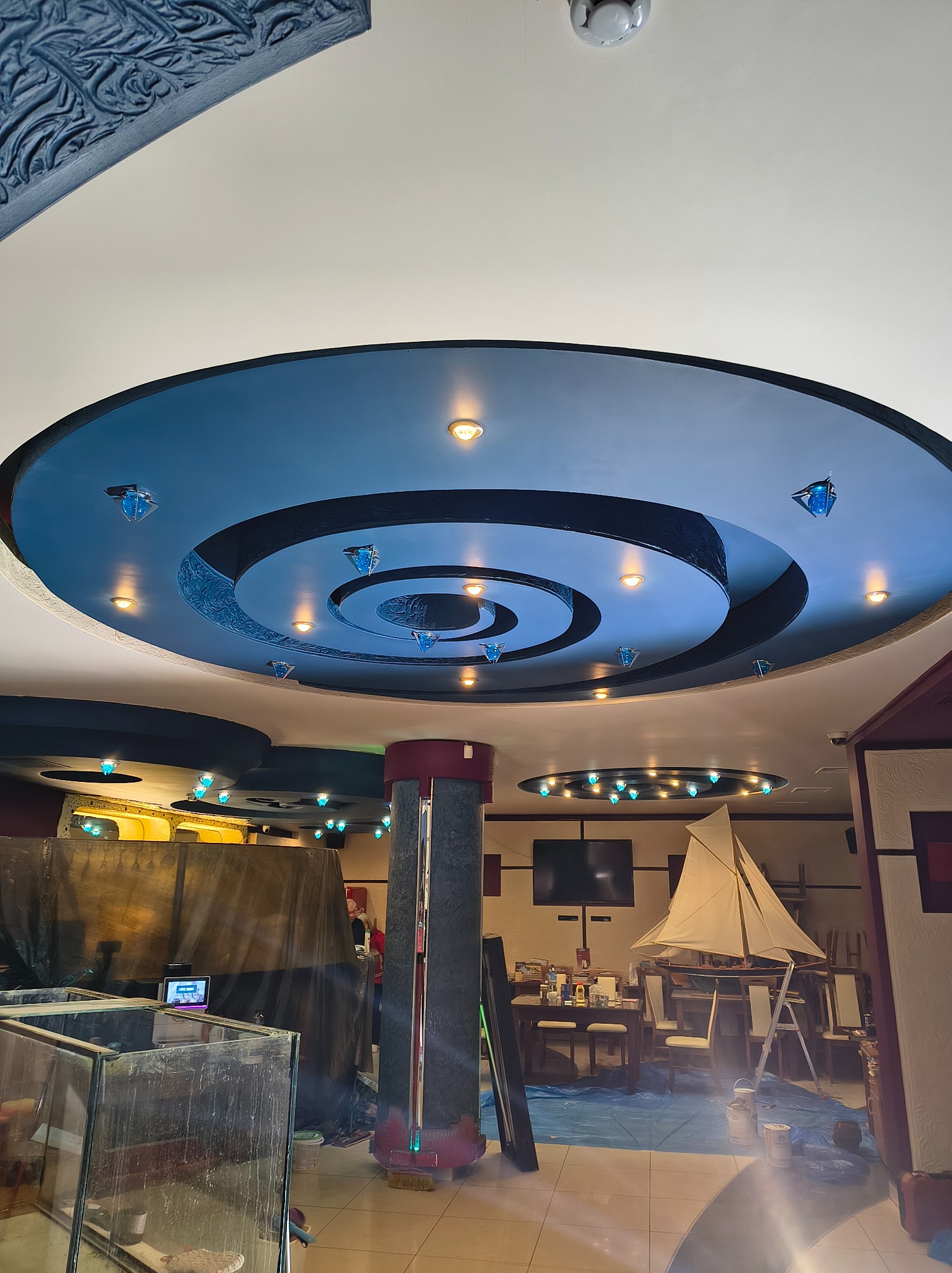 Dekoracyjny sufit z niebieskimi okręgami i oświetleniem punktowym w trakcie malowania, widoczne zabezpieczenia podłogi i elementy wyposażenia wnętrza, w tle model żaglowca.