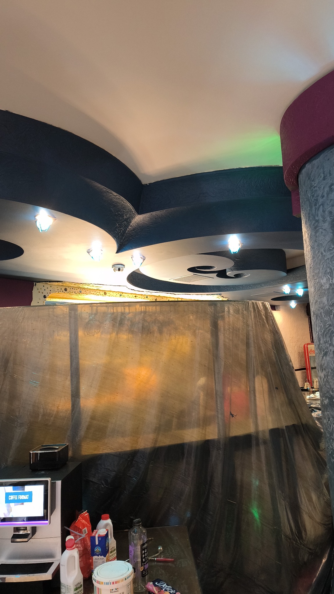 Przygotowania do malowania sufitu w biurze: zakryte meble folią ochronną, widoczny pojemnik z farbą i automat do kawy w tle, dekoracyjny sufit z oświetleniem.