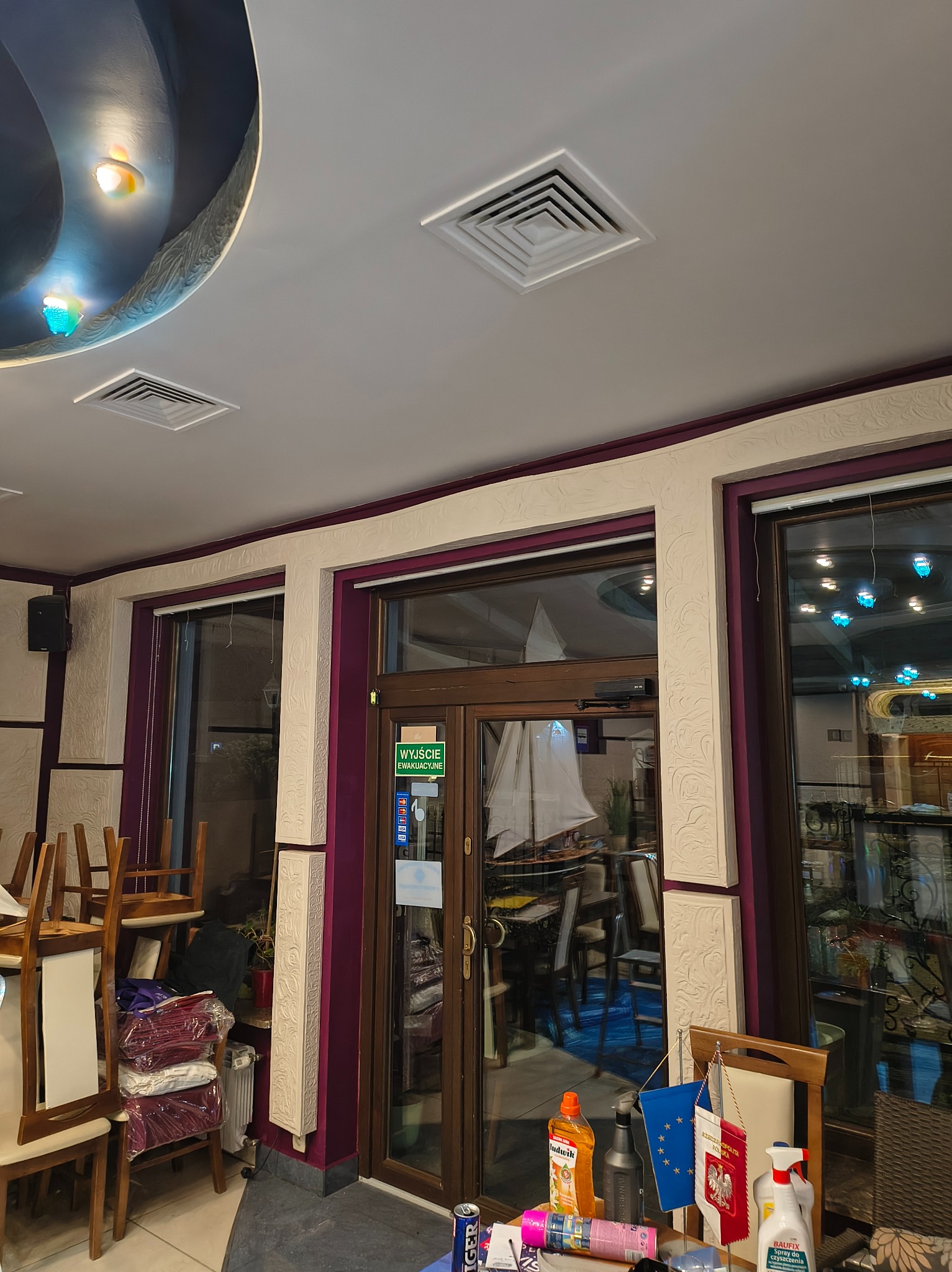 Wnętrze lokalu gastronomicznego z widocznym sufitem z dekoracyjnym oświetleniem, kratkami wentylacyjnymi, oknami z dekoracyjnymi obramowaniami i częściowo widocznym wyposażeniem, w tym krzesłami...