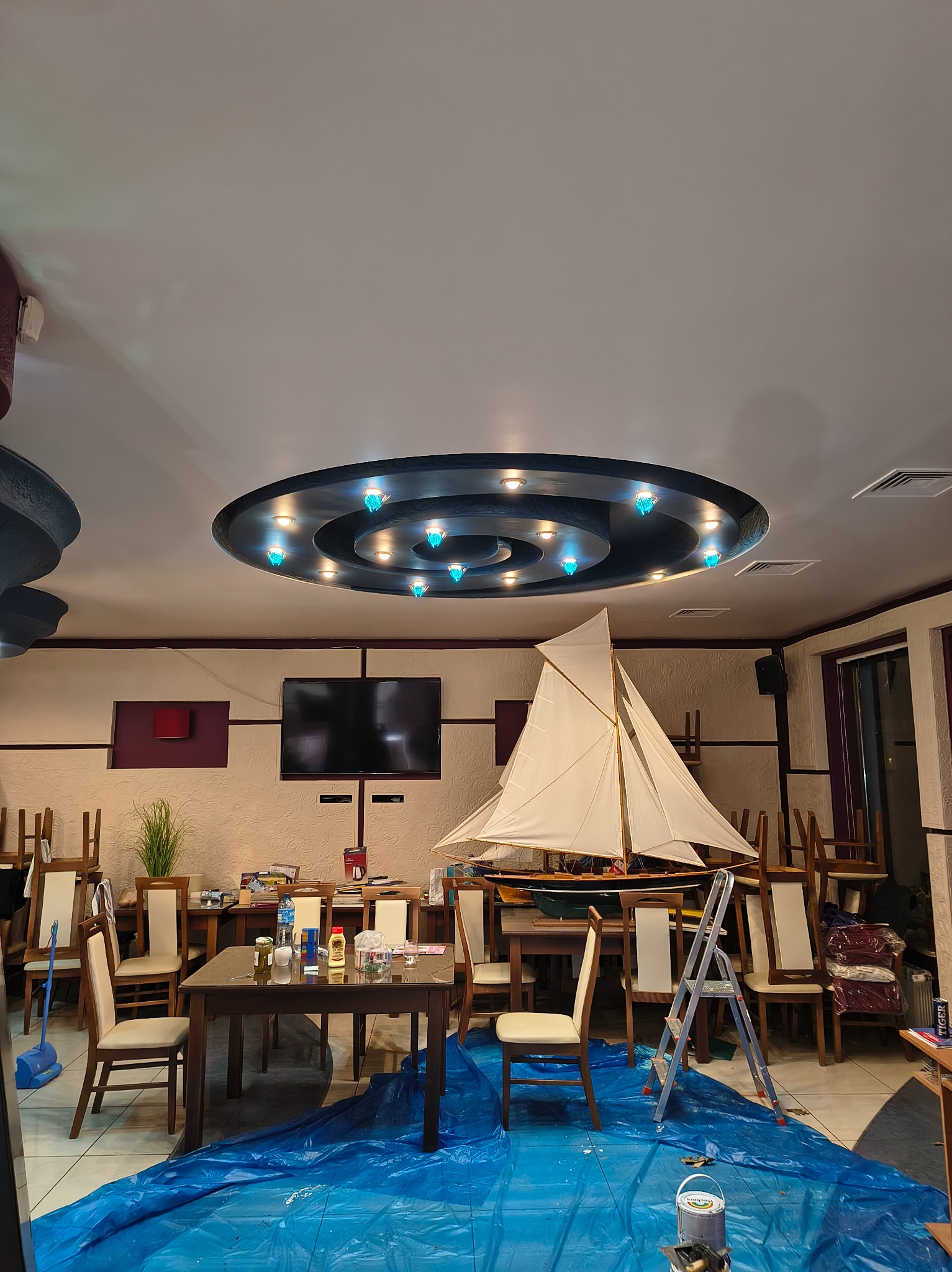 Przygotowania do malowania w restauracji: meble zabezpieczone folią, drabina, model żaglowca na stole, odwrócone krzesła na stołach, ozdobne oświetlenie sufitowe.