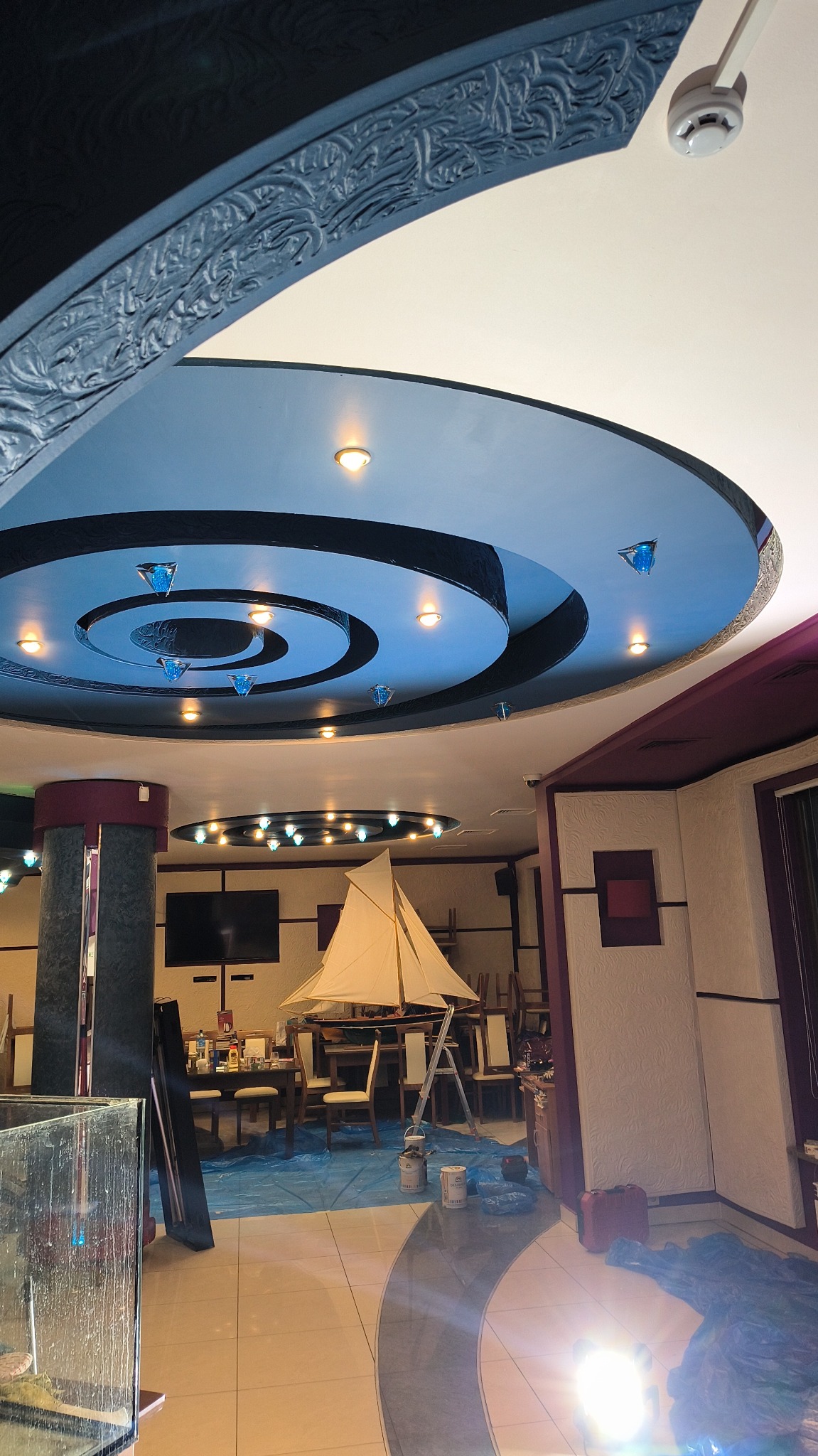 Malowanie sufitu w restauracji z ozdobnymi elementami i oświetleniem, widoczne zabezpieczenia podłogi folią malarską i drabina.