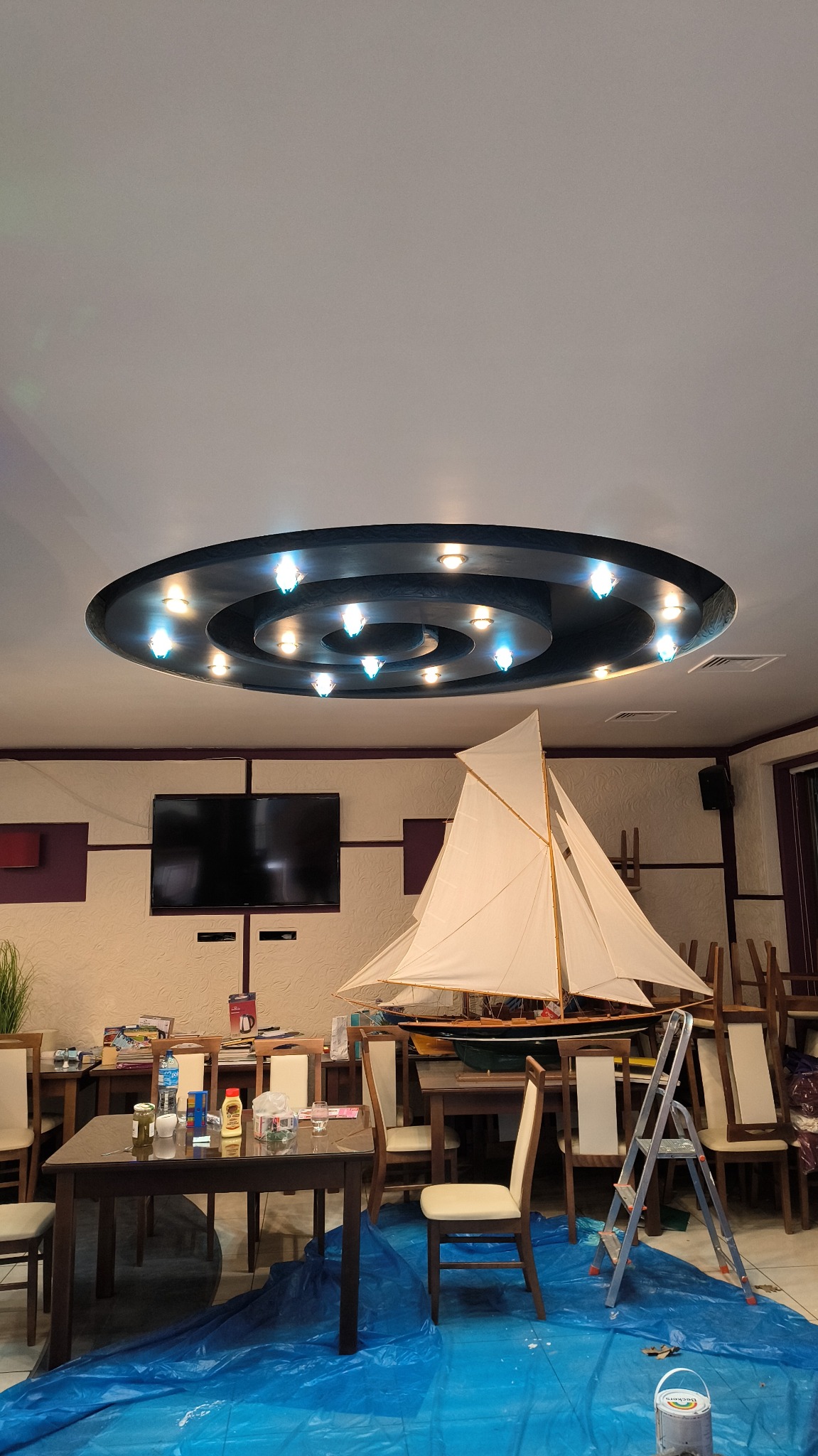 Pokój z zabezpieczonym folią podłożem, stołami i krzesłami, model żaglówki i drabina sugerują prace remontowe w toku, prawdopodobnie malowanie sufitu z okrągłą lampą.