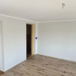 Wnętrze odświeżonego pokoju z widoczną częścią kuchni, świeżo pomalowane ściany na jasny odcień szarości, drewnopodobne panele na podłodze, wystające przewody elektryczne z sufitu.