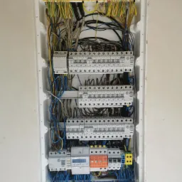 Rozdzielnica elektryczna z zainstalowanymi bezpiecznikami, licznikiem energii i przewodami w kolorach żółto-zielonym, niebieskim i szarym, zamontowana w białej obudowie w ścianie.