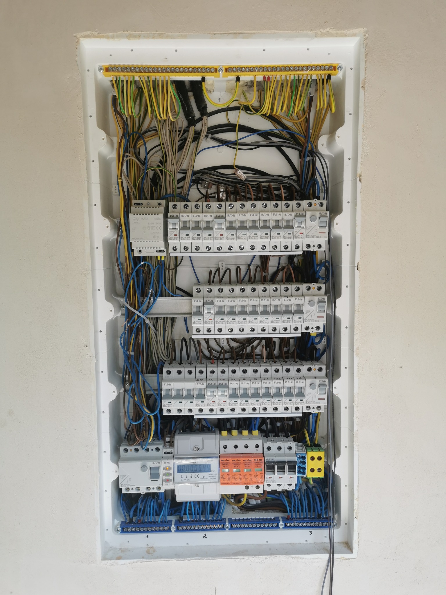 Rozdzielnica elektryczna z zainstalowanymi bezpiecznikami, licznikiem energii i przewodami w kolorach żółto-zielonym, niebieskim i szarym, zamontowana w białej obudowie w ścianie.
