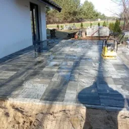 Ułożona kostka brukowa w odcieniach szarości, mokra od deszczu, z żółtą zagęszczarką w tle, widoczne przygotowane podłoże i palety z kostką.