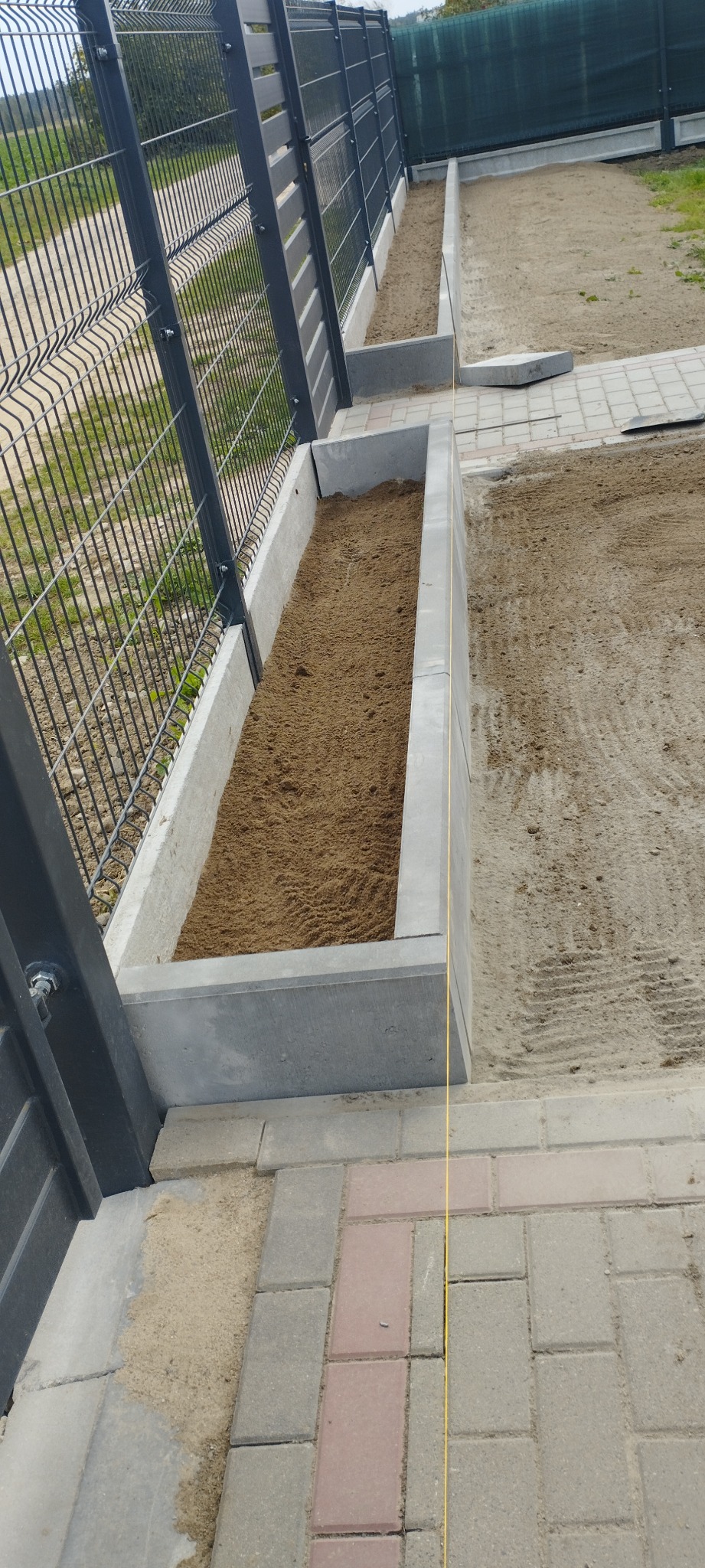 Przygotowanie terenu pod rabaty kwiatowe z obrzeżami z betonowych elementów wzdłuż ogrodzenia panelowego z siatki. Żółta linka wyznacza linię prostej krawędzi rabaty.