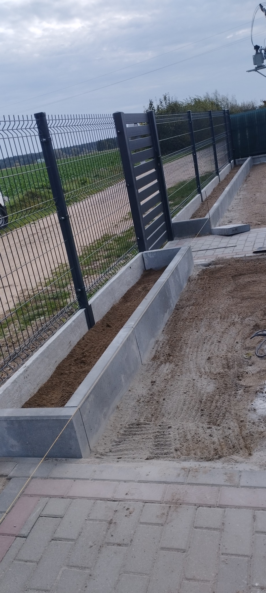 Montaż ogrodzenia panelowego z elementami betonowymi oraz układanie kostki brukowej wzdłuż ogrodzenia, widoczna ziemia w betonowych korytach.