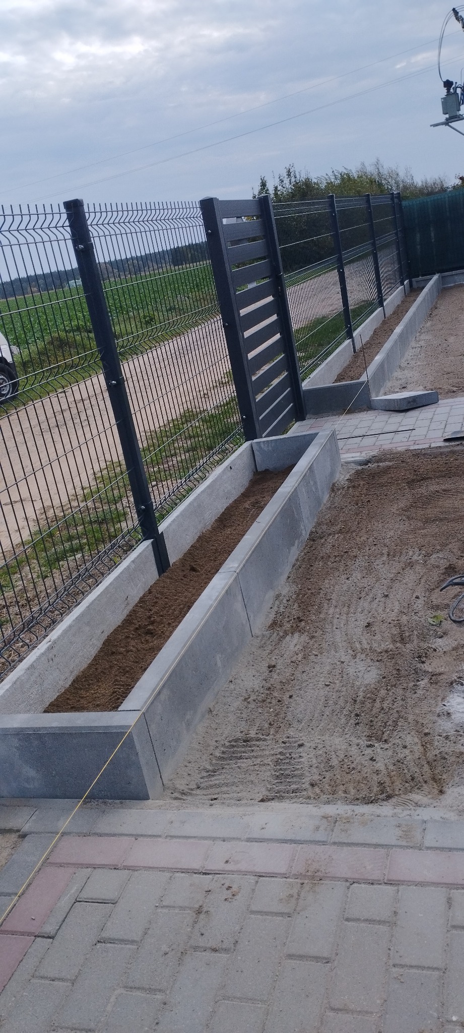 Montaż czarnego ogrodzenia panelowego z elementami metalowymi i betonowymi korytami wypełnionymi ziemią, obok ułożona kostka brukowa.