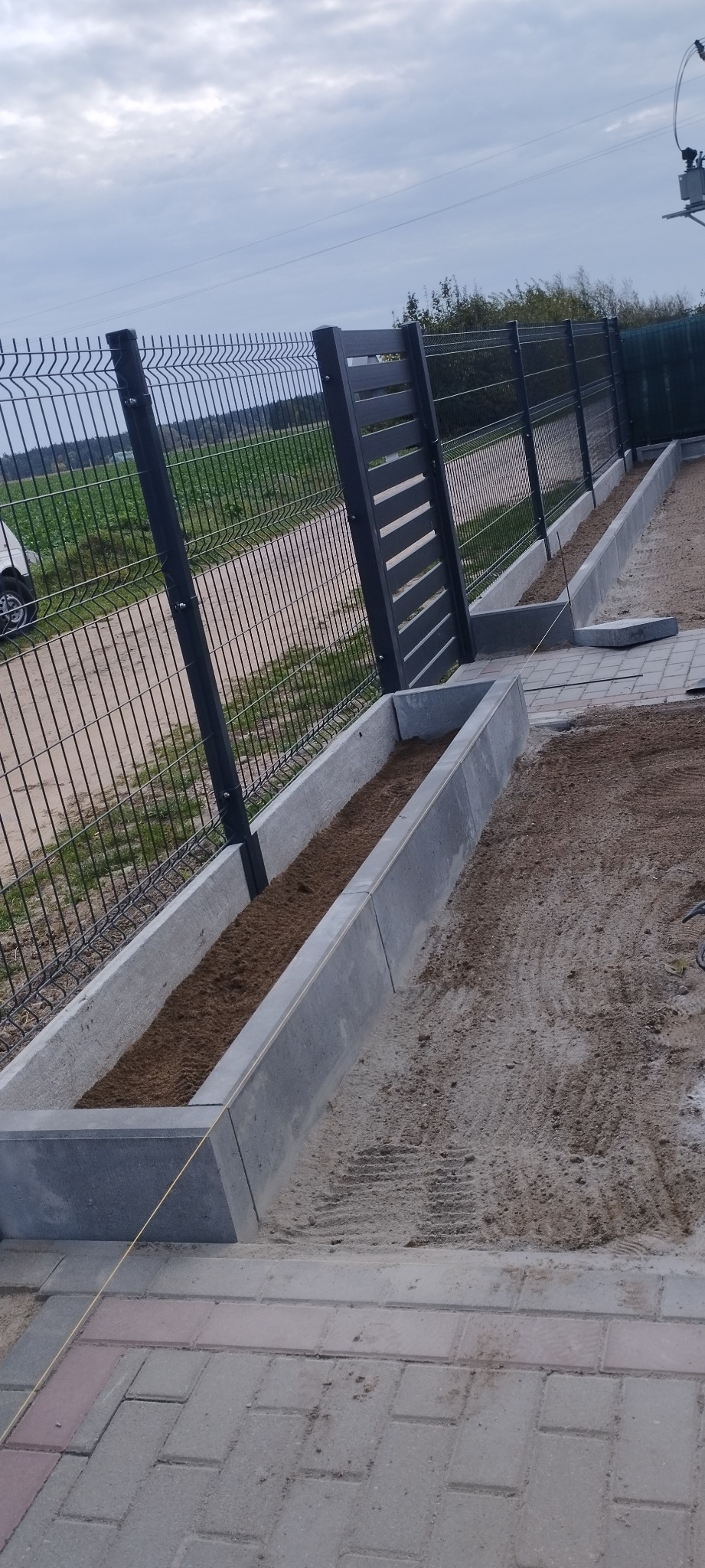 Montaż ogrodzenia panelowego z betonową podmurówką i układanie kostki brukowej w trakcie realizacji, widoczna linka wyznaczająca linię bruku.