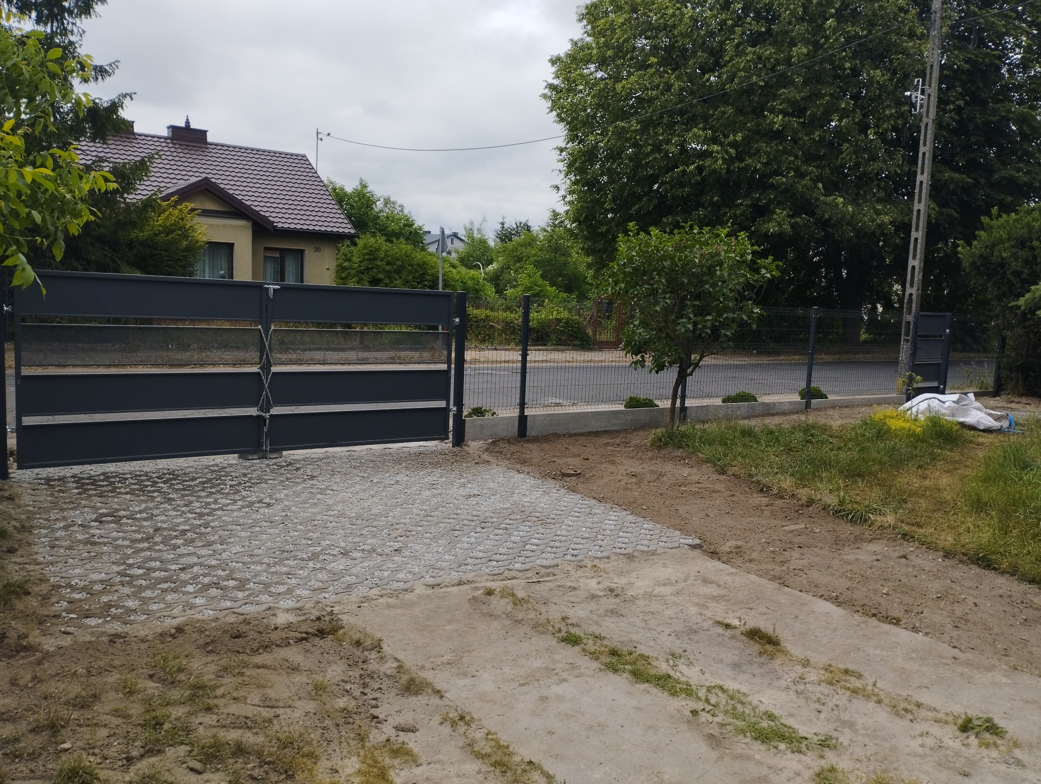 Nowoczesna posesja z szarą, przesuwną bramą i ażurową kostką brukową na podjeździe, uzupełniona siatką ogrodzeniową i młodymi drzewkami w tle.