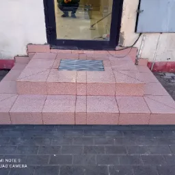 Wykończone schody wejściowe do budynku, wyłożone r&oacute;żowymi płytkami, z kratką odpływową u g&oacute;ry, widoczne odbicie osoby w drzwiach.