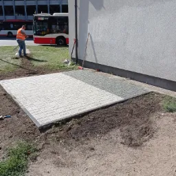 Układanie kostki brukowej na nowym chodniku, widoczny pracownik porządkujący teren grabiami, w tle autobus i budynek parkingu.