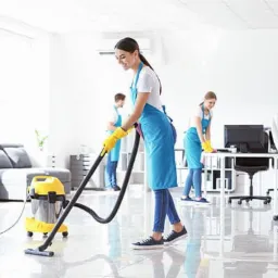 Kobieta w niebieskim fartuchu odkurza błyszczącą podłogę biura żółtym odkurzaczem przemysłowym; w tle dwie inne osoby sprzątają biurka.