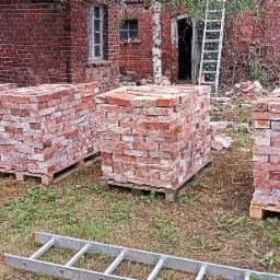 Ułożone na paletach cegły z odzysku przed budynkiem w trakcie rozbiórki, widoczna drabina i zarośnięte otoczenie.