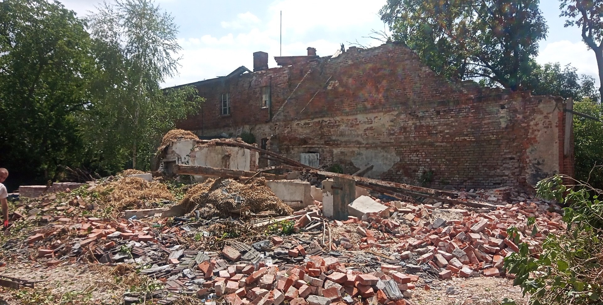 Częściowo zburzony budynek z czerwonej cegły, gruzowisko i fragmenty konstrukcji na pierwszym planie, widoczne drzewa w tle.