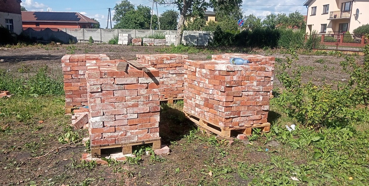 Stosy czerwonej cegły na drewnianych paletach, składowane na ziemi z widoczną zielenią, w tle budynki i ogrodzenie.