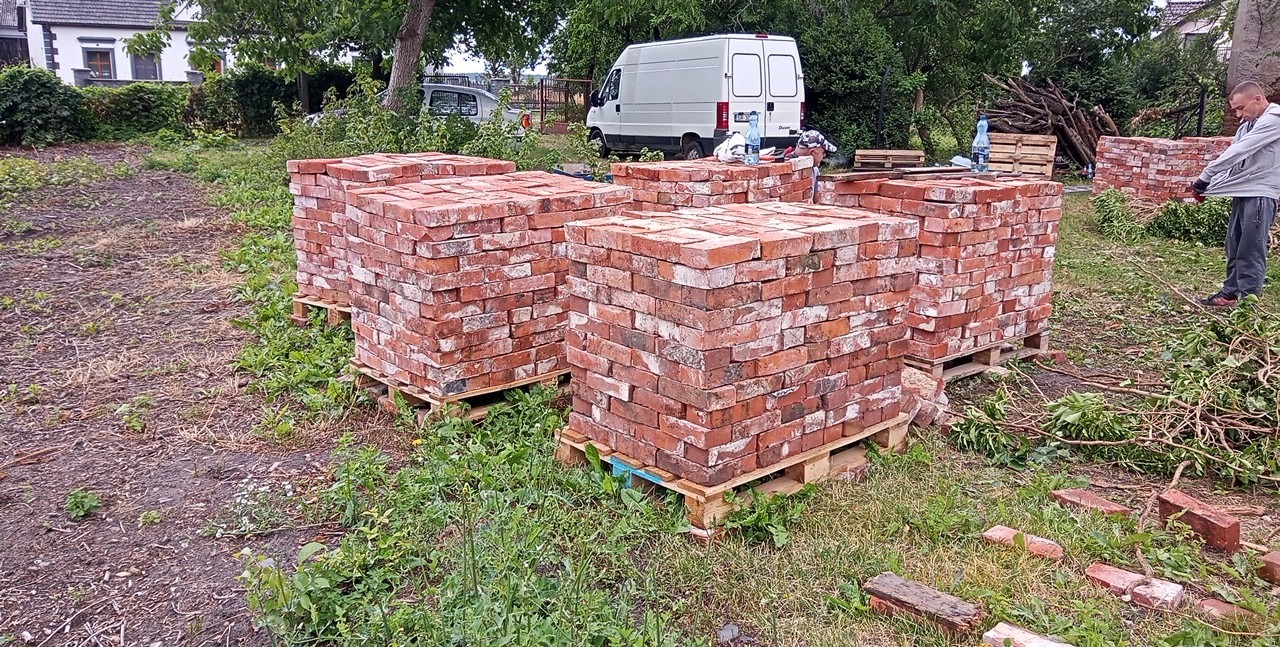 Ułożone na paletach cegły rozbiórkowe w ogrodzie z widocznym busem w tle, mężczyzna w jasnej bluzie i sterta gałęzi.