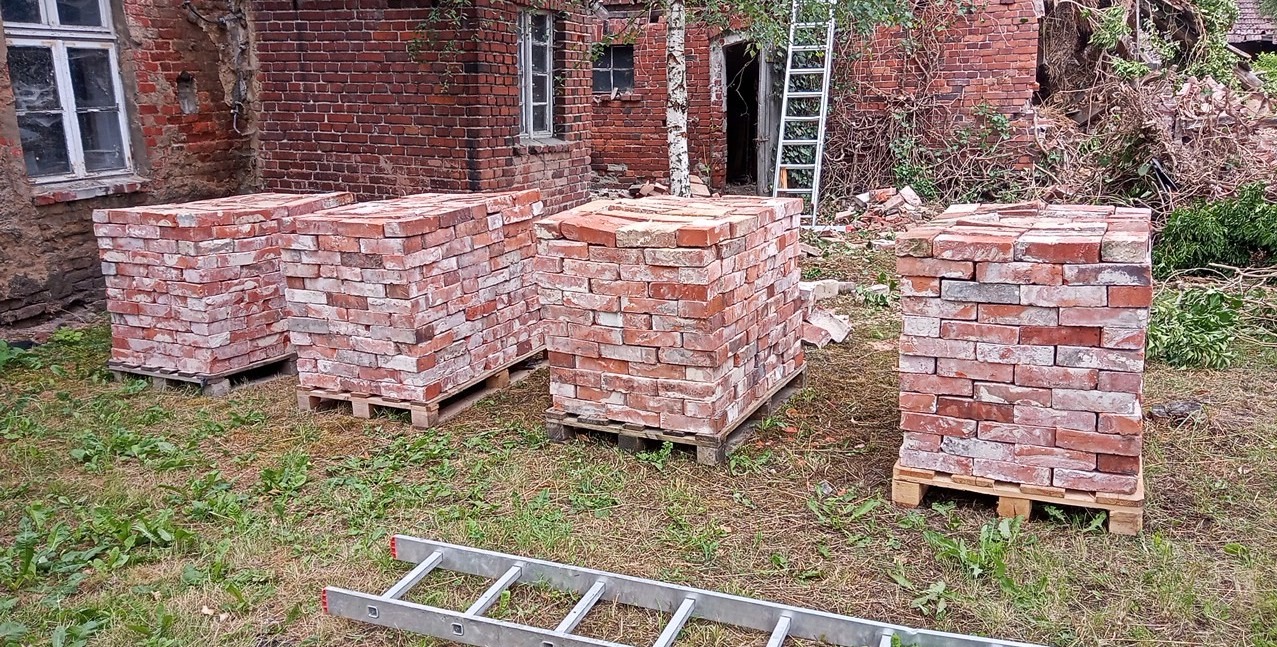 Ułożone na paletach cegły z odzysku przed budynkiem w trakcie rozbiórki, widoczna drabina i zarośnięte otoczenie.