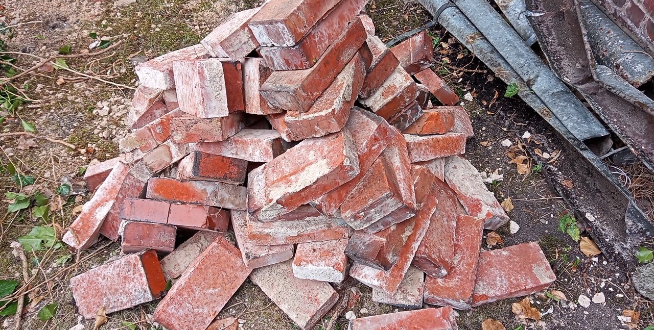 Stos czerwonych, używanych cegieł z resztkami zaprawy, leżący na ziemi obok zardzewiałych, metalowych rur.