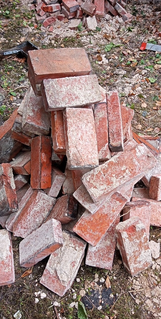 Stos czerwonych, używanych cegieł z resztkami zaprawy, ułożonych na ziemi z fragmentami liści i trawy.