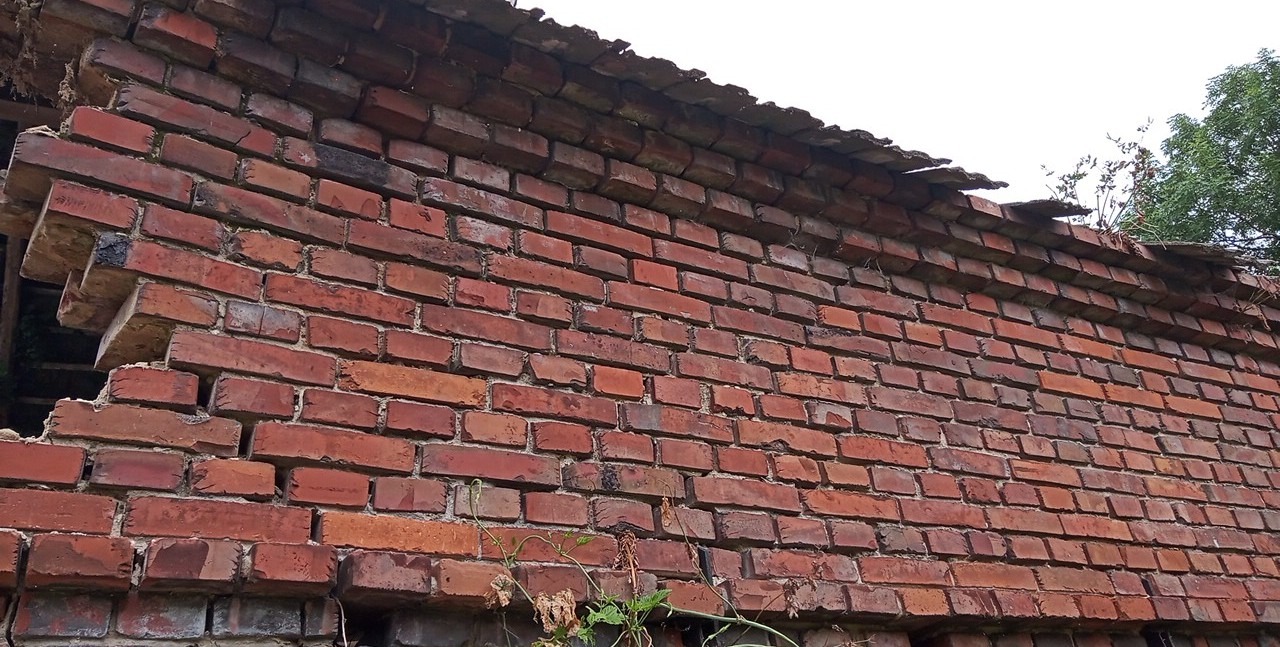 Uszkodzony mur z czerwonej cegły, fragment budynku w trakcie rozbiórki, widoczne ubytki i wystające cegły, zarośnięty roślinnością u podstawy.