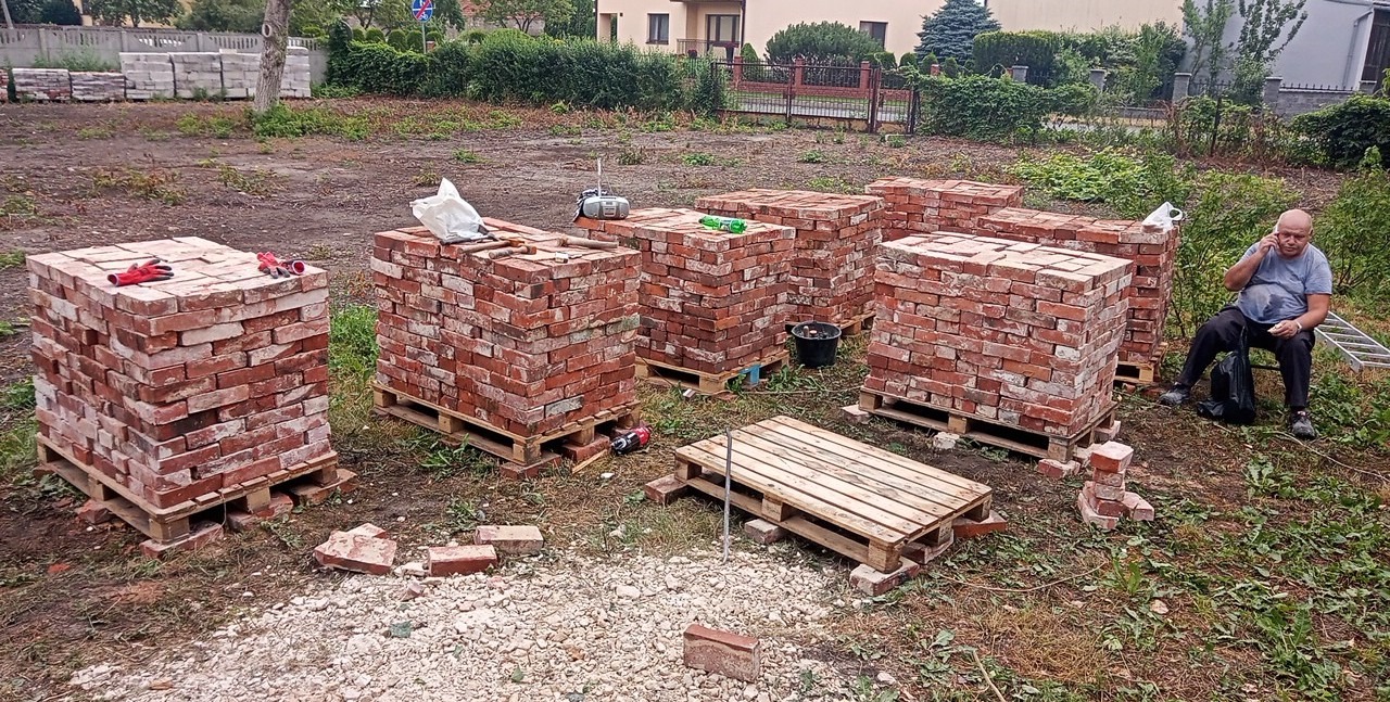 Stosy czerwonej cegły na paletach, przygotowane do rozbiórki lub budowy, mężczyzna siedzi obok, rozmawiając przez telefon.