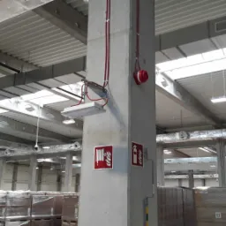 Oświetlenie i system przeciwpożarowy w hali magazynowej: lampa LED zamontowana na betonowym słupie, czerwone przewody, sygnalizator pożaru oraz znaki bezpieczeństwa.
